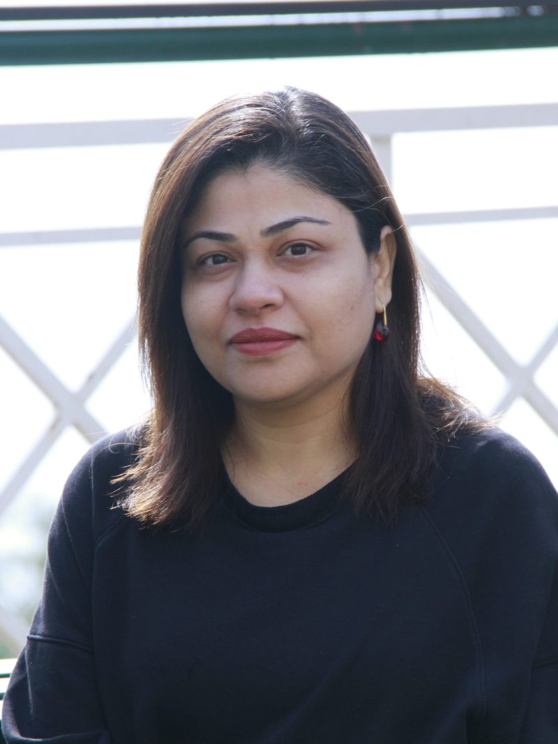 A profile photo of Leena Ahmed. She is wearing a dark blue shirt, a gold and red reaching dangles from the ear that's visible as her brown hair is tucked behind it. 