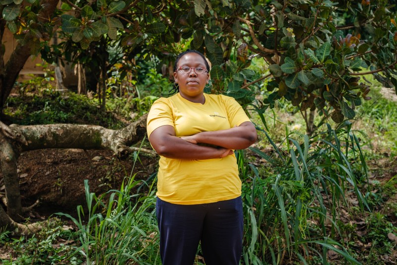 Omosefe stands in front of greenery of grass, bushes and trees. She is wearing light auburn colored eye glasses, thin gold hoops in each ear, a yellow Sightsavers t-shirt and blue trousers. Her arms are crossed as she looks into the camera with a serious look. 